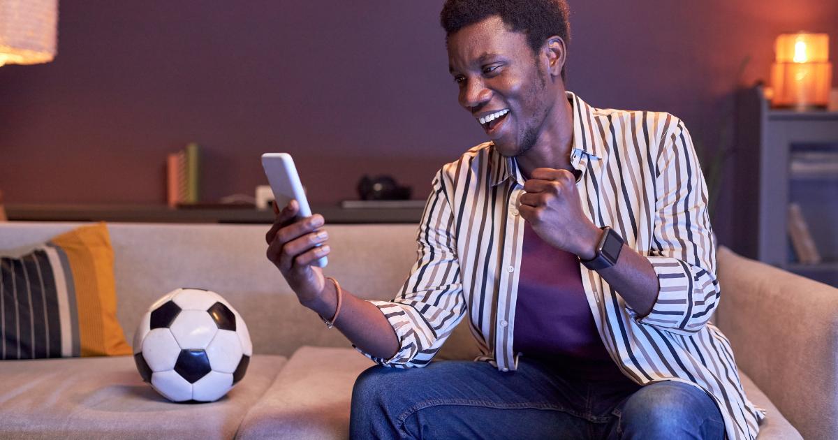 Man in his living room watching his betting app on his phone next to a soccer ball.